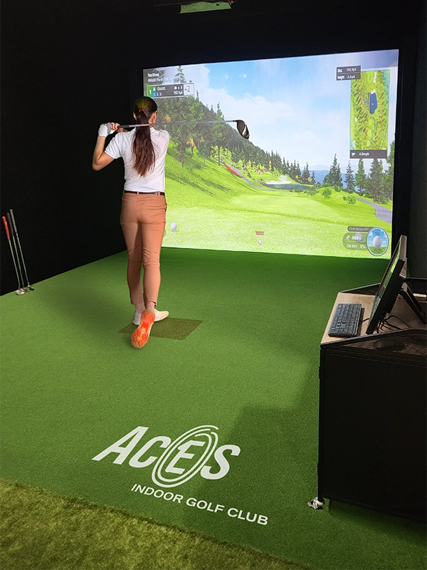 A female golfer preparing to swing in a virtual golf environment at Aces Indoor Golf Club, standing in front of a large screen projecting a golf course.