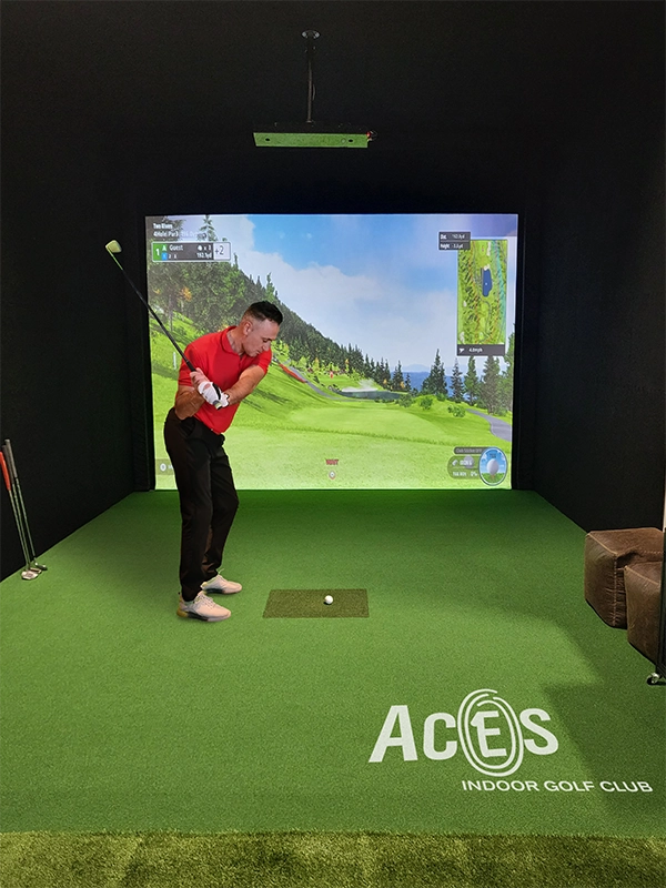 A male golfer mid-swing in a virtual golf setting at Aces Indoor Golf Club, aiming towards a large screen displaying a golf course.